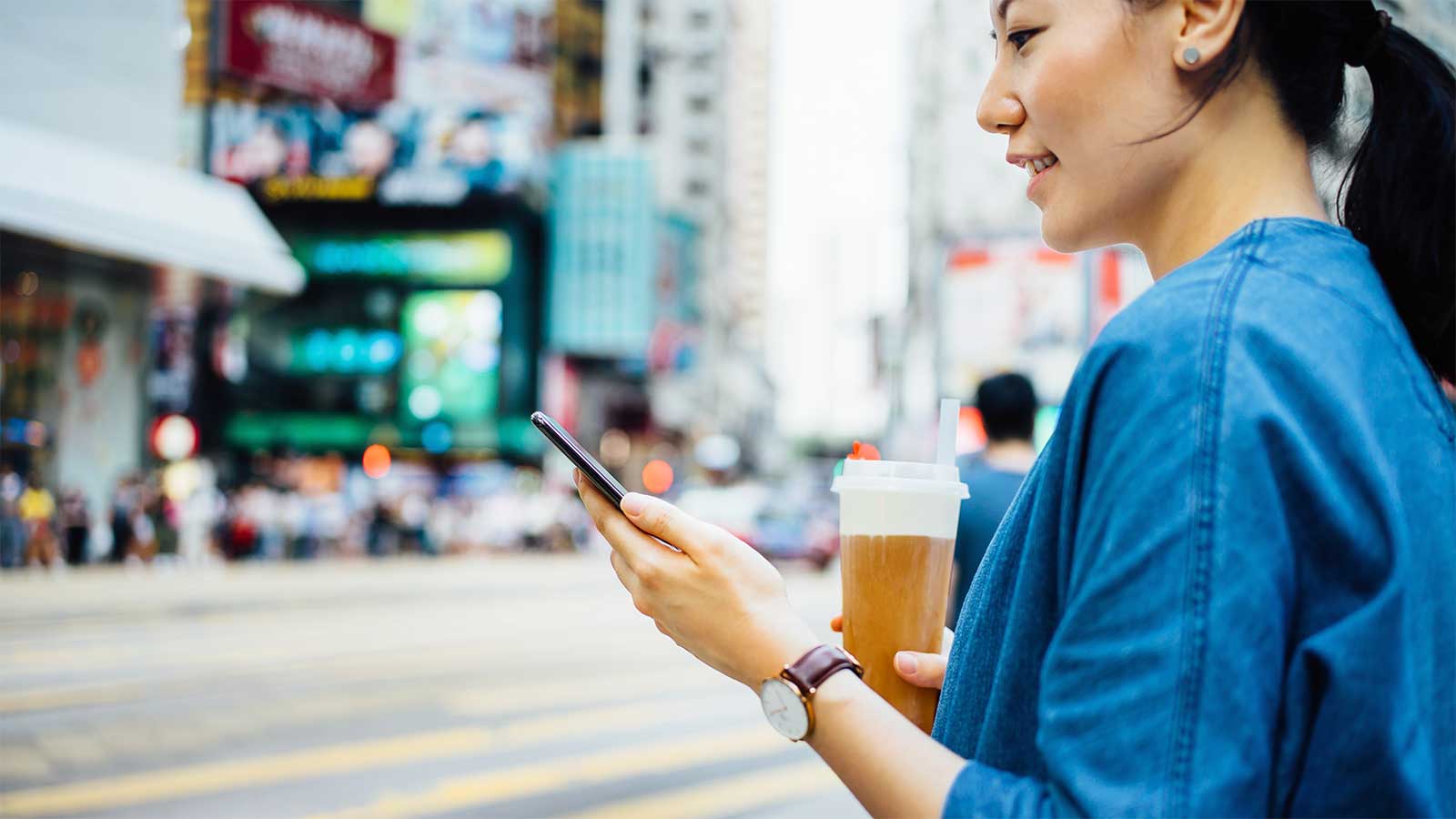 Gros plan d’une femme avec une boisson glacée dans une main et un smartphone dans l’autre, attendant devant un passage piéton de pouvoir traverser une rue animée