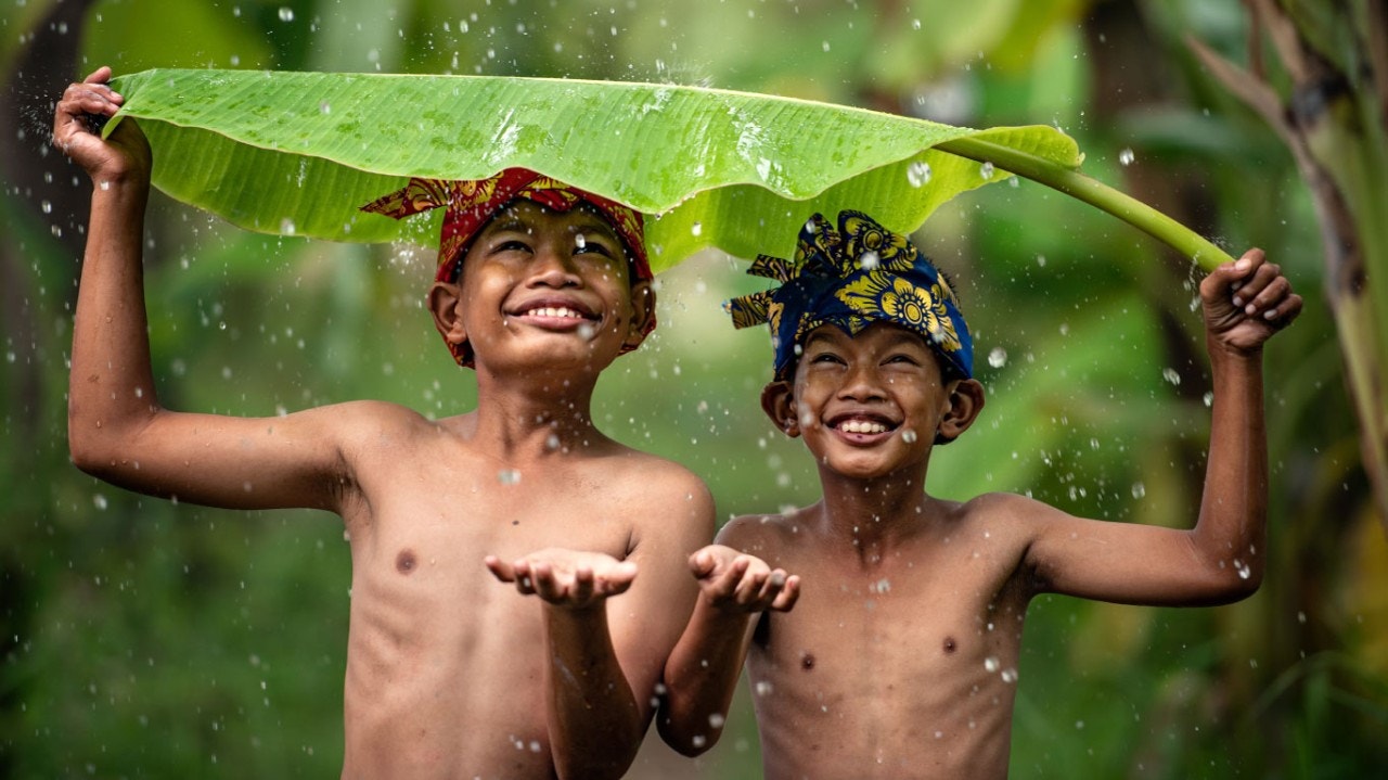 Des garçons asiatiques riant dehors sous la pluie, l’un d’eux tenant une énorme feuille de bananier