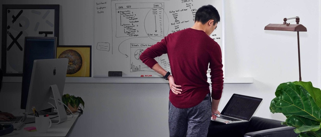 Back view of a man standing in front of a whiteboard using a laptop