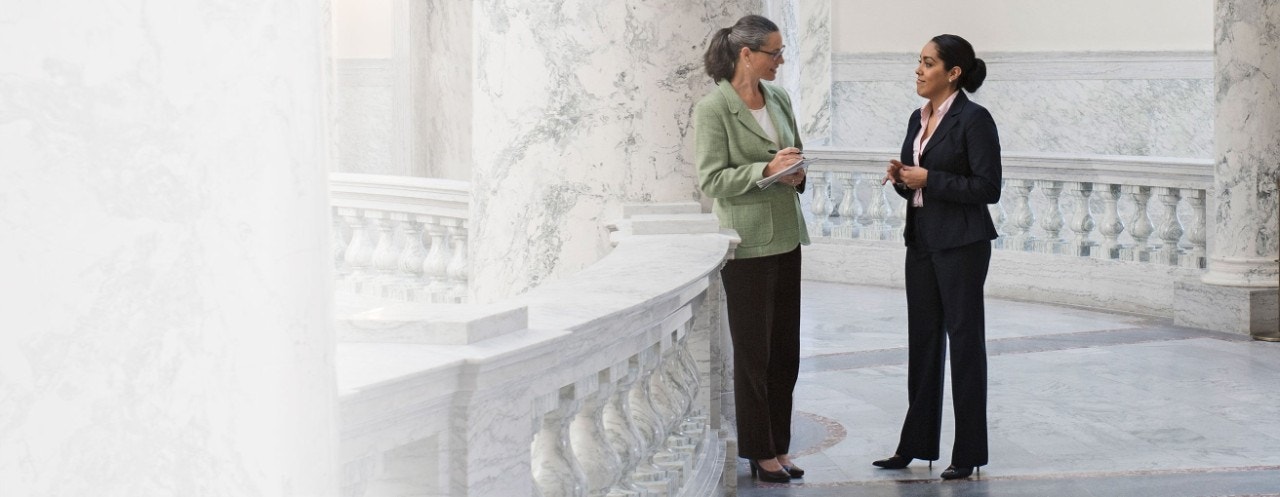 Two people discussing while standing in the hallway of a white marble building