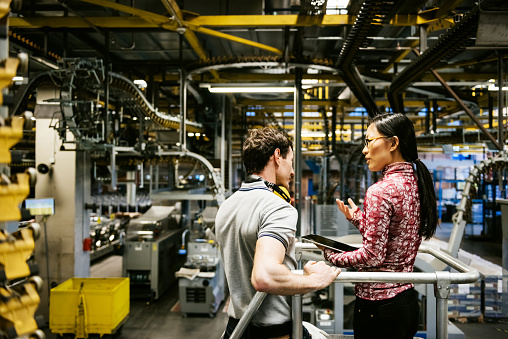 Person holding tablet in discussion with coworker in industrial setting surrounded by pipes and equipment