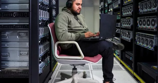 Pessoa sentada em uma cadeira em um local de armazenamento de dados usando fones de ouvido e trabalhando em um laptop