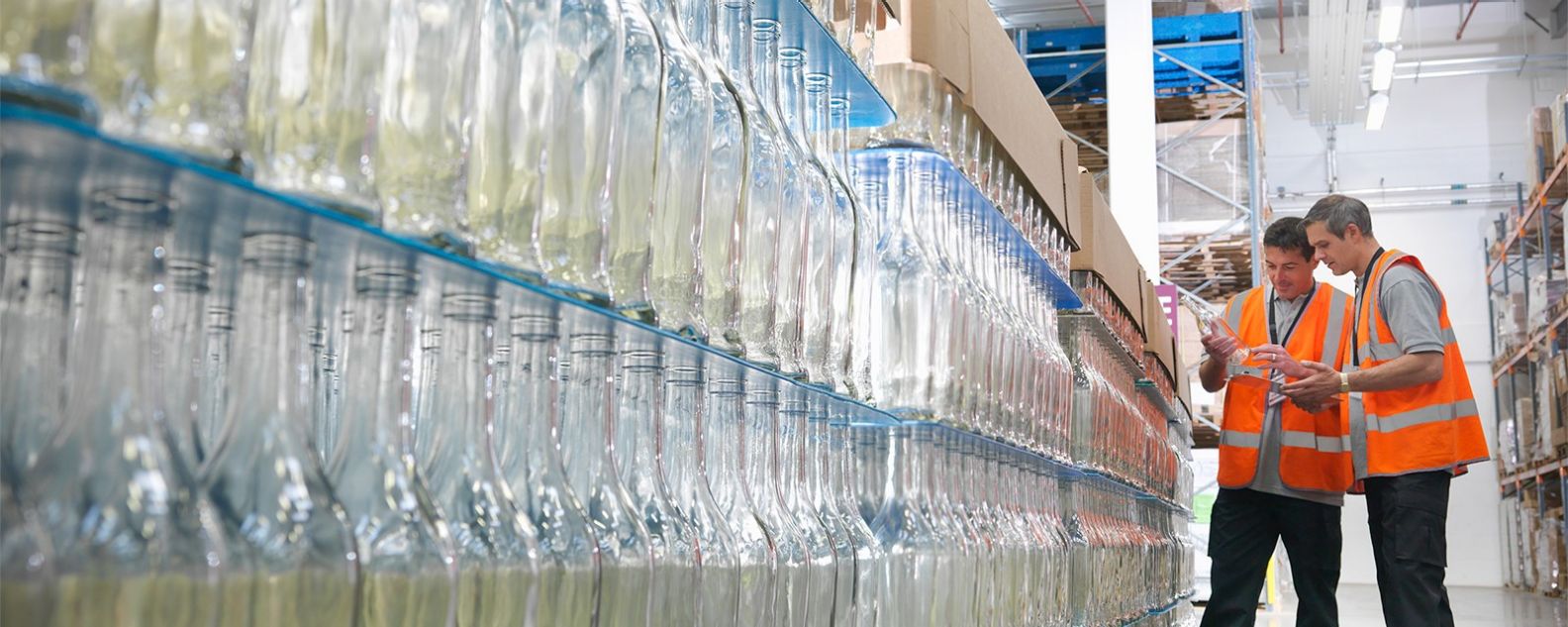 Dos trabajadores de una fábrica con chalecos naranjas examinan estanterías de botellas transparentes