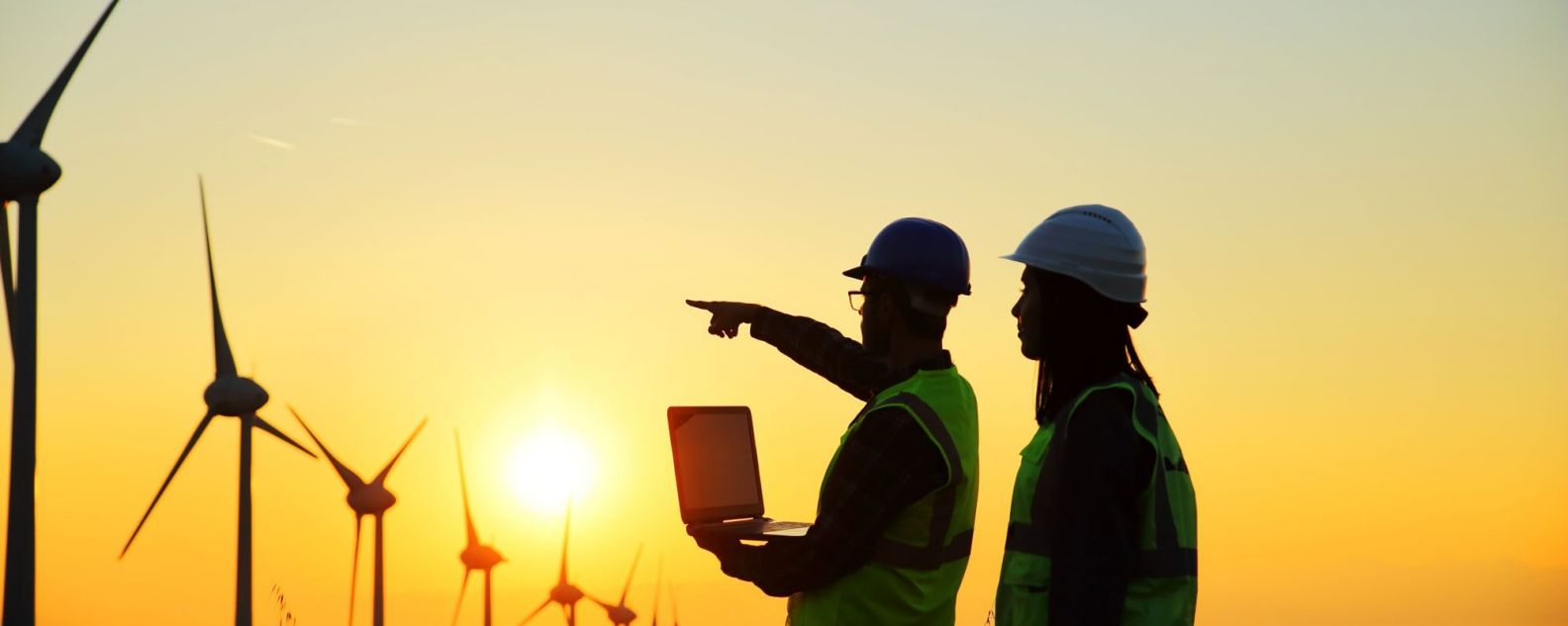 Two workers, one pointing to wind machines with a sunset background