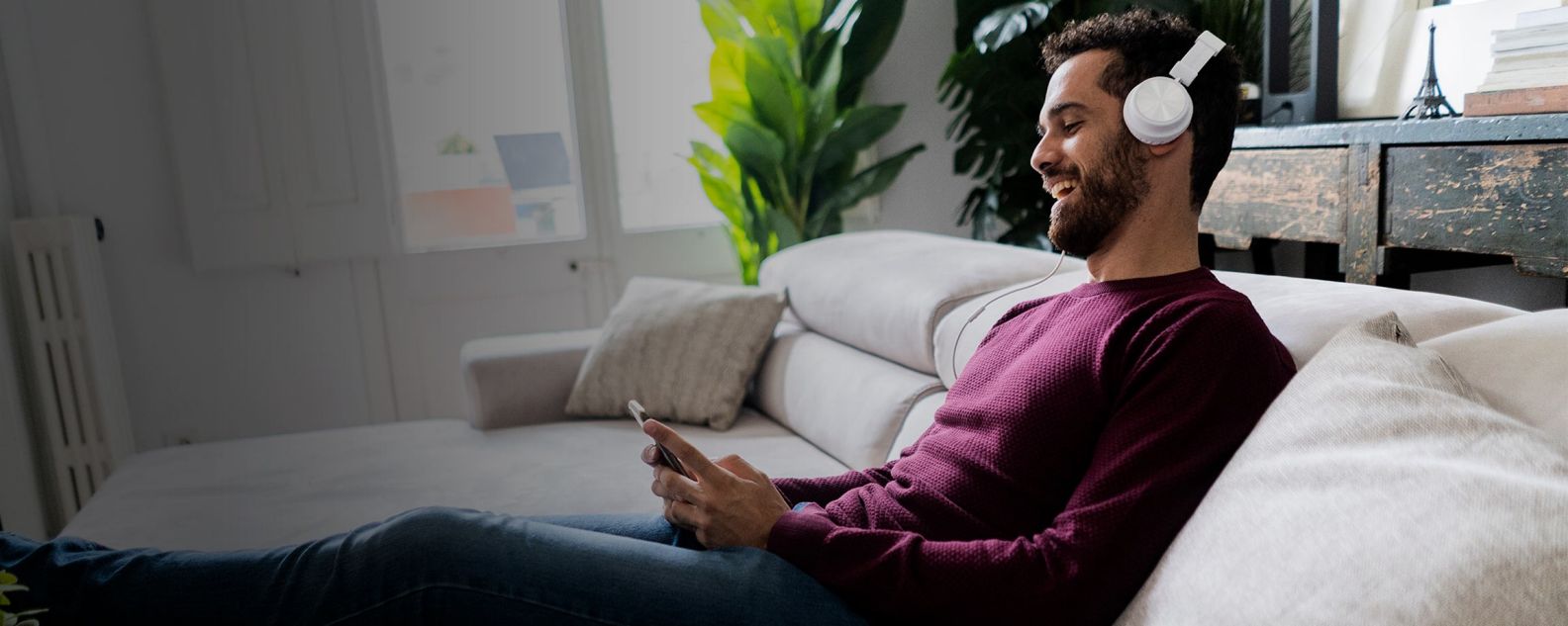 Homem rindo sentado no sofá com celular e fones de ouvido