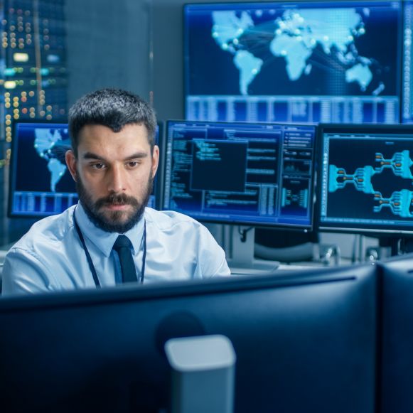 Lavoratore presso un PC desktop, con monitor sullo sfondo che mostrano schermate sulla sicurezza