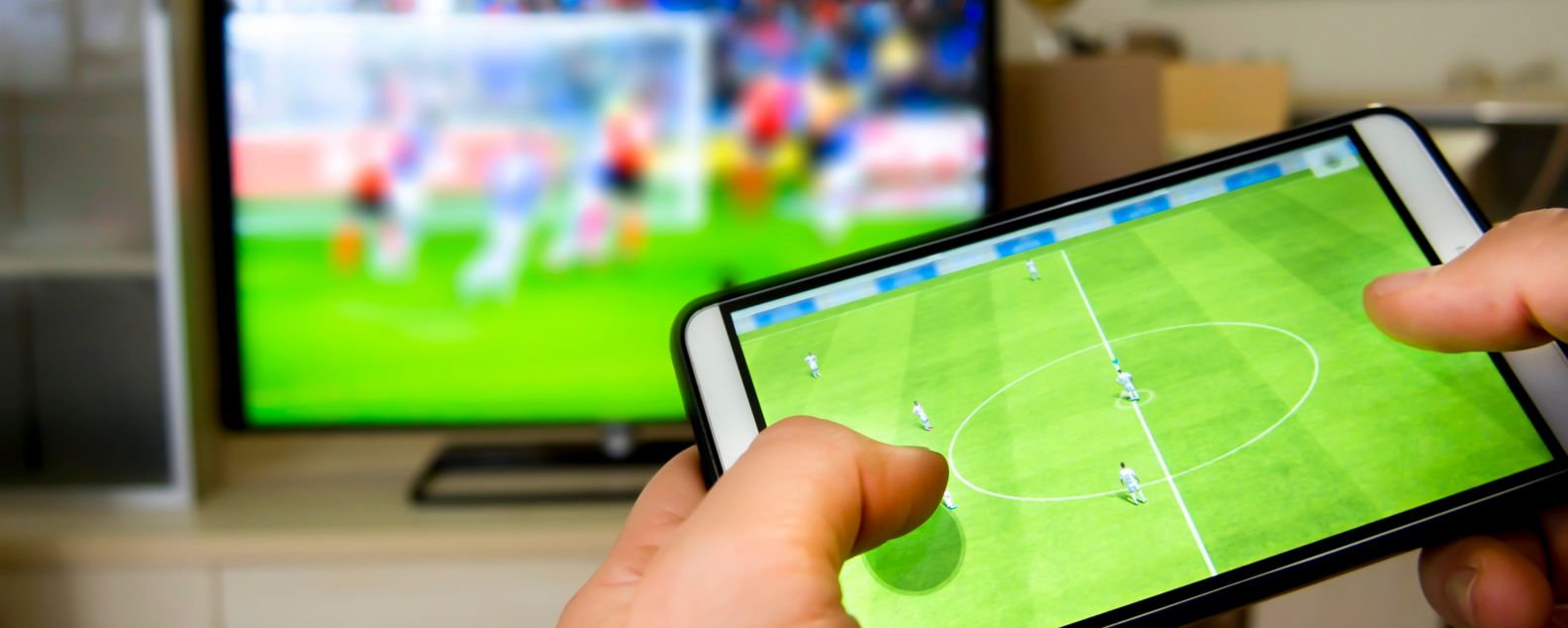 Persona interactuando con un partido de fútbol en un dispositivo móvil mientras ve un partido en la televisión