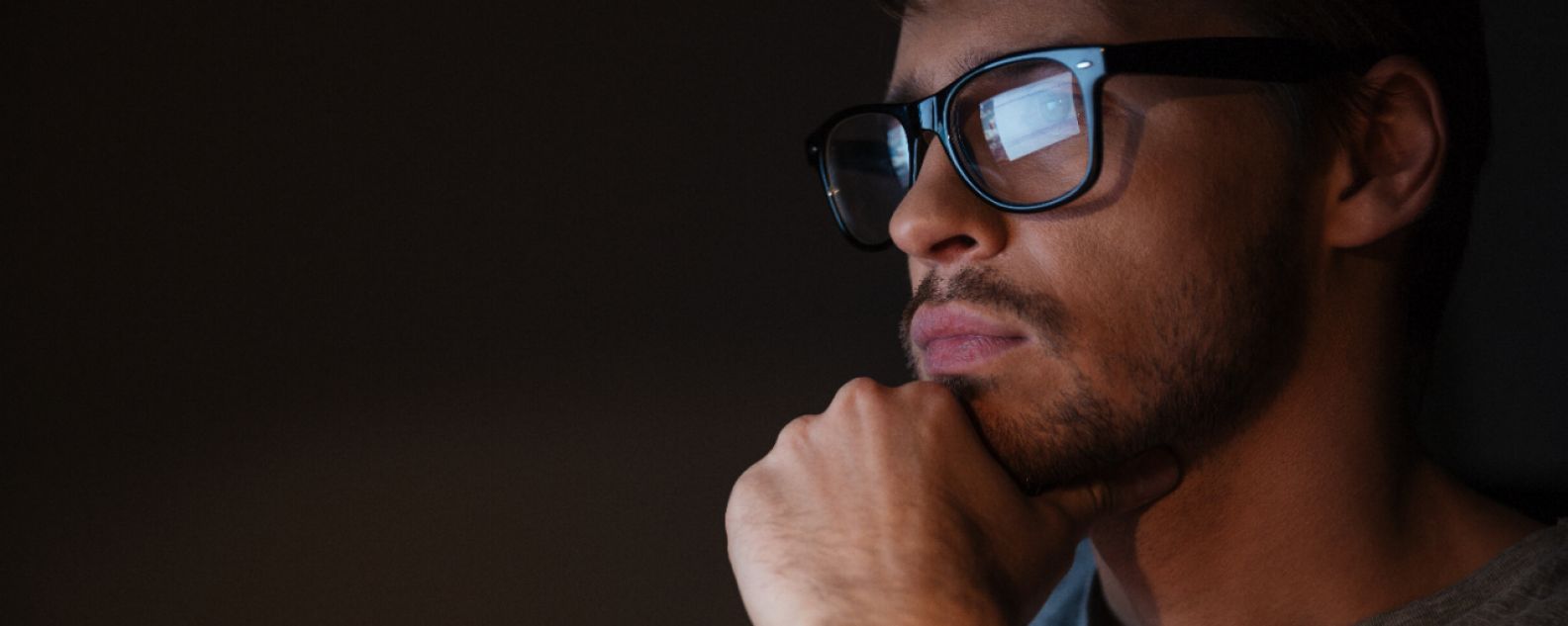 Detalhe de um homem pensativo, com a tela do computador refletida em seus óculos