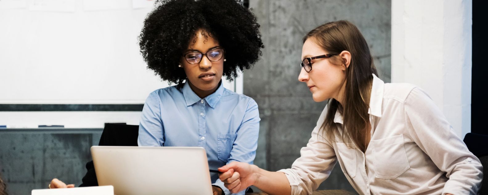一名非裔和一名白人年轻女子正在现代化办公室出席商务会议。他们通过文件和笔记本电脑讨论一些事情，其中一人指着电脑屏幕。两人都戴着眼镜，背景中的饼图清晰可见。