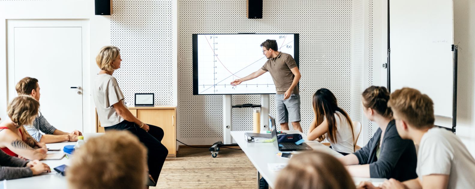 Seorang mahasiswa memberikan presentasi di depan kelas pada sesi seminar dan menggunakan monitor besar sebagai alat bantu visual