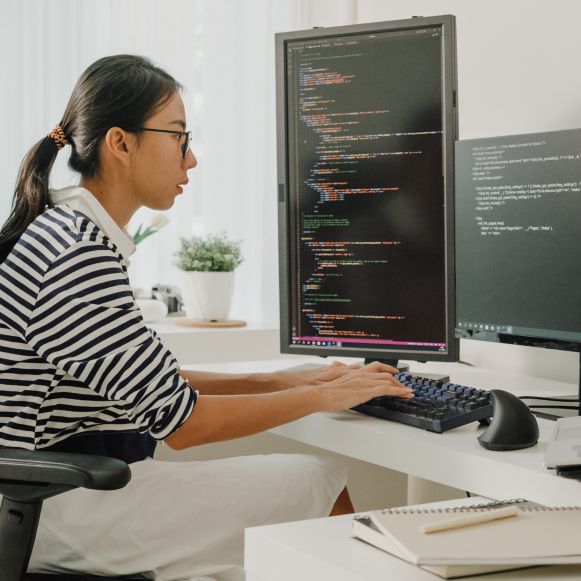 안경을 쓴 채 여러 개의 화면이 있는 책상에 앉아 키보드로 코드를 입력하고 있는 젊은 아시아 여성 소프트웨어 개발자