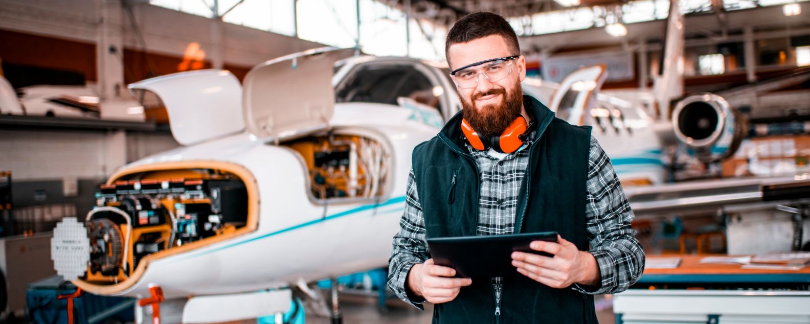 Flugzeugingenieur lächelt und blickt in die Kamera, während er ein digitales Tablet in der Hand hält und ein Flugzeug repariert und wartet