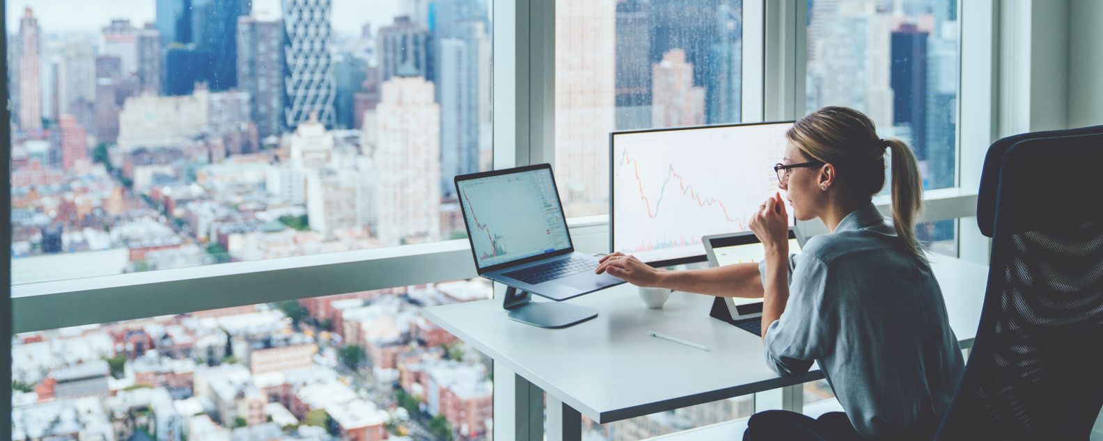 Vista posteriore di una persona seduta al computer desktop dell'ufficio di un grattacielo panoramico con grafici finanziari e statistiche sul monitor