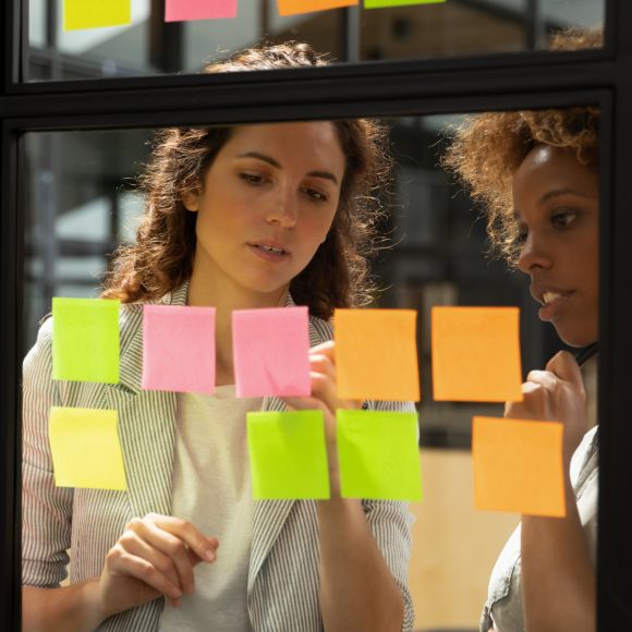 Compañeras de trabajo escribiendo en notas adhesivas de colores pegadas a una ventana de oficina