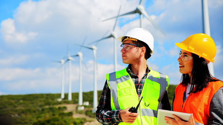 Two employees using a tablet device at open field with multiple wind turbines in the back