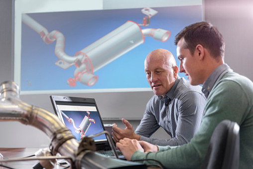 Two workers reviewing equipment design on laptop while image is projected on large screen behind them