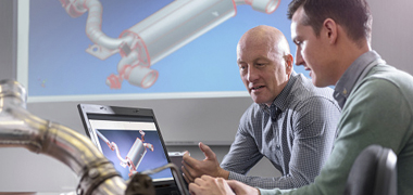 Two workers reviewing equipment design on laptop while image is projected on large screen behind them