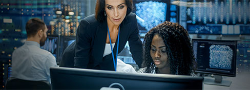 Two professionals working on a computer with another individual in the background on a computer