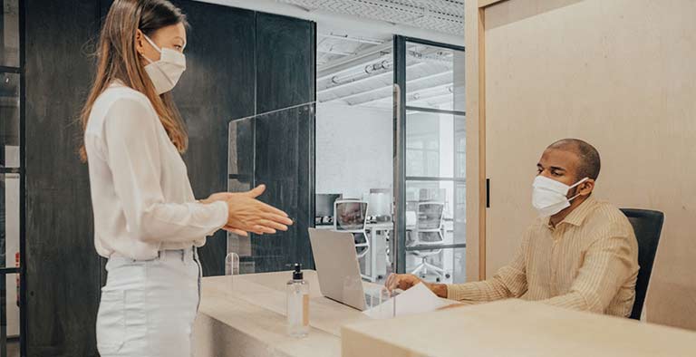 An employee cleans her hands with sanitizer, while her coworker is positioned behind a glass partition.