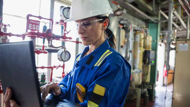 Woman standing in uniform and helmet working on her notebook in oil and gas company