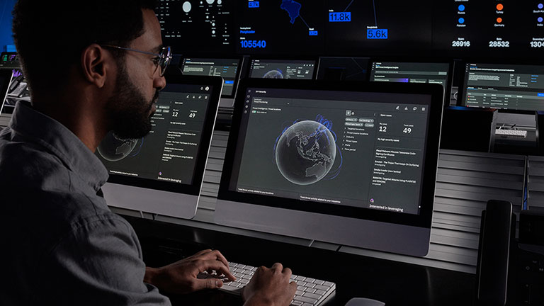 A man working on a work station in a control center with status screens in the background.