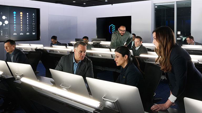 People working at work stations in a control center with status screens in the background.