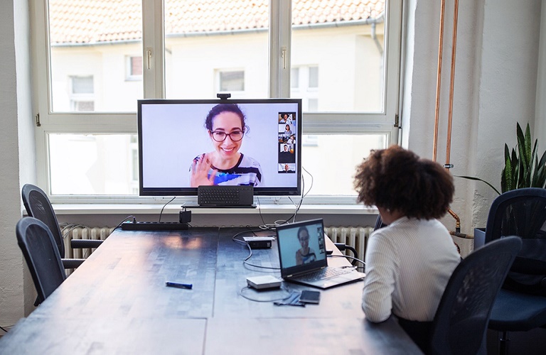 A woman in front of a monitor showing a virtual team meeting.