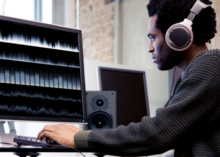 Man wearing headphones typing on keyboard and looking at computer monitor displaying five separate sound waves