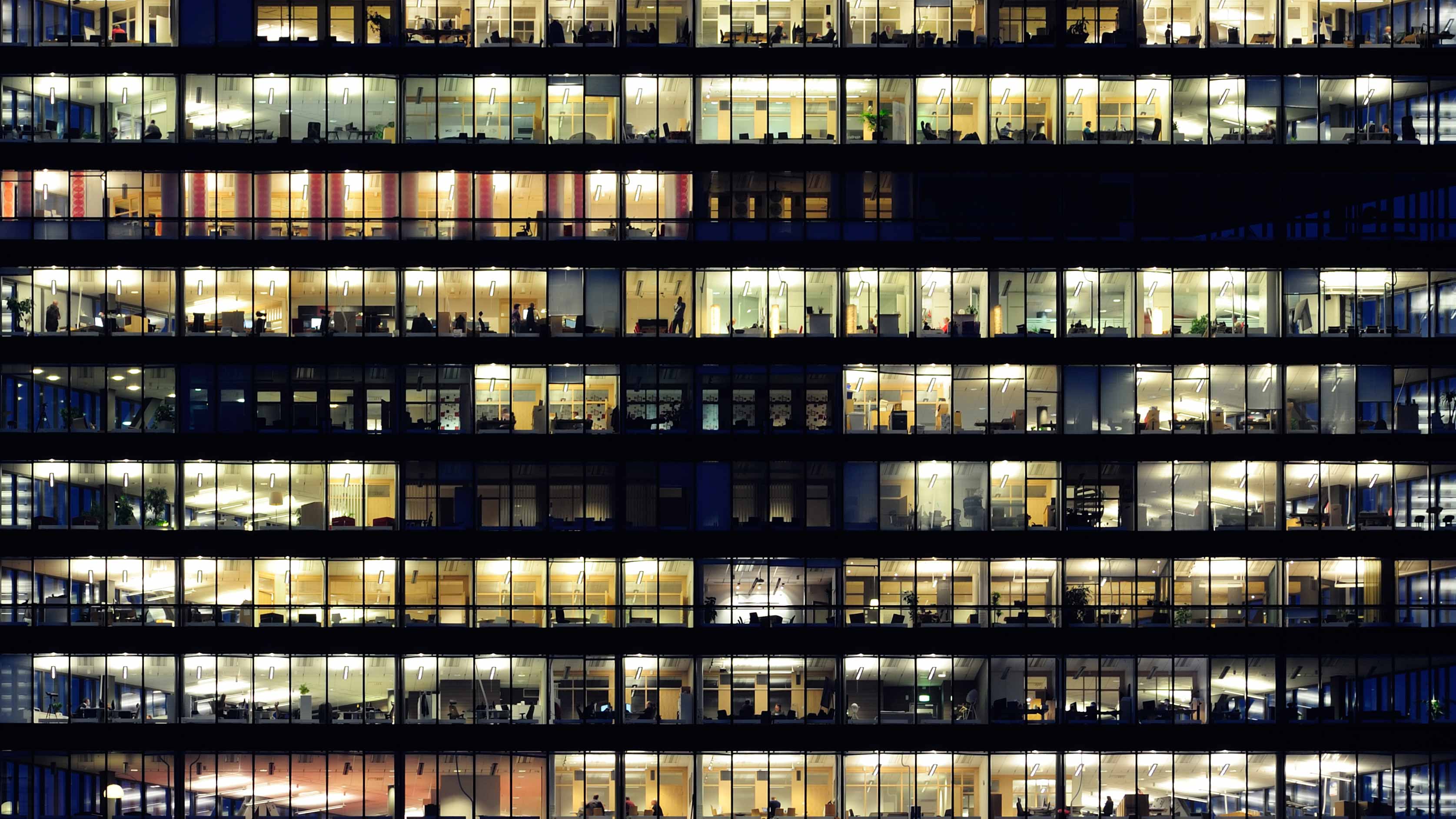 Uma imagem ampla da fachada de um prédio comercial à noite, com metade das janelas dos escritórios iluminadas.