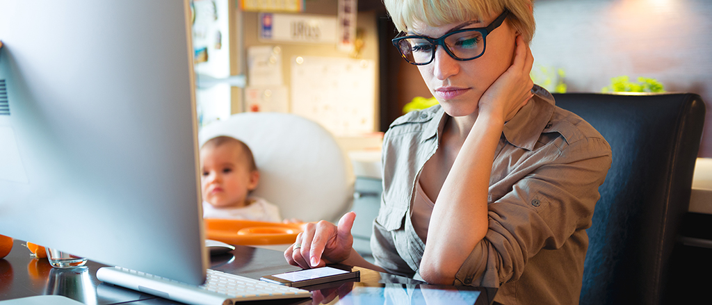 A woman looking at a phone and an iPad with a baby on a highchair in the background