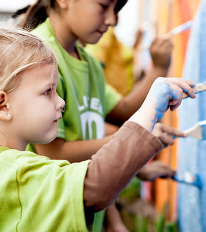 Pre-school aged girl in a green t-shirt and with blue paint on her hands works on a school mural with other youth volunteers