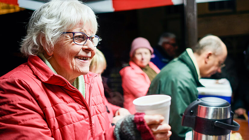 Smiling senior citizen woman wearing a red parka offers up a warm cup of coffee and a healthy dose of cheer at a food kitchen