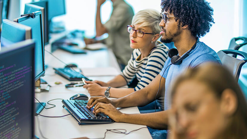 A young developer in a blue t-shirt and with headphones around his neck reviews code with his female colleague