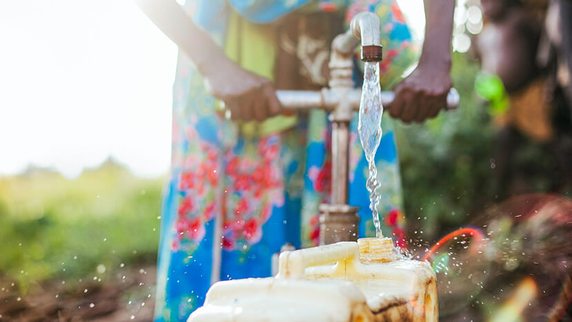As villagers look on, a black woman in a floral dress pumps water into a large container from a community well in Africa