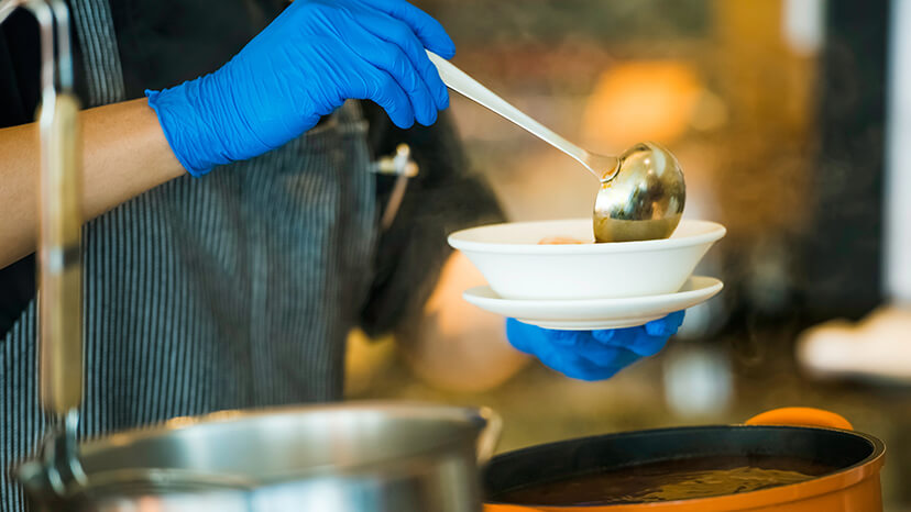 Close-up of a soup kitchen volunteer wearing protective blue latex gloves and an apron asthey ladle soup into a white bowl