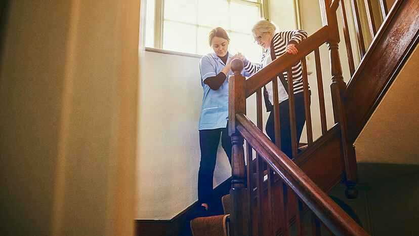 A young caregiver holds the hand of an elderly woman as they carefully descend a flight of wooden stairs to start the day