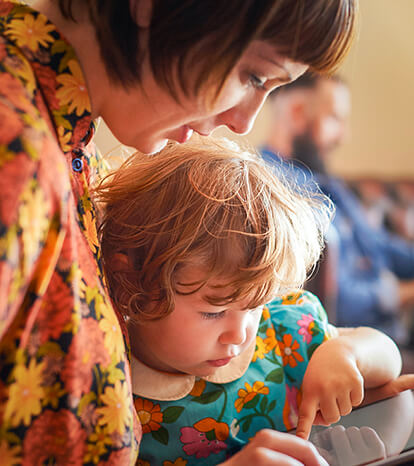 A young mom in a floral dress shares a story with her red-haired infant daughter as the girl’s father reads in the background