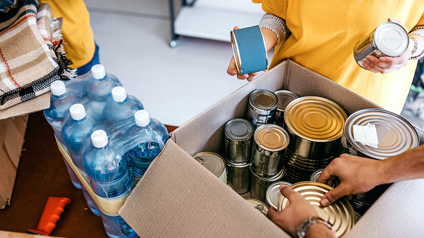 Food bank volunteers in yellow t-shirts pack a cardboard box with canned goods, bottled water and blankets for those in need