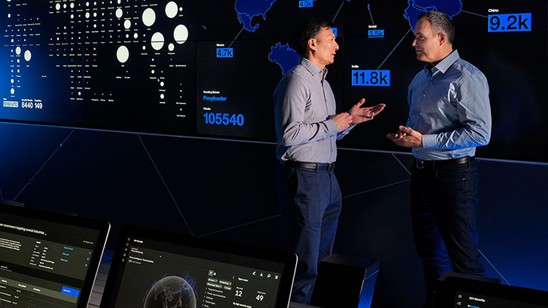 Two men talking in a control room in front a large status screen with a map of the world.